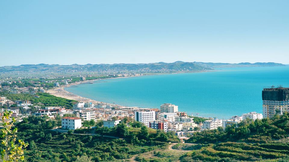 A coastal cityscape with a beach, buildings, and mountains in the background.
