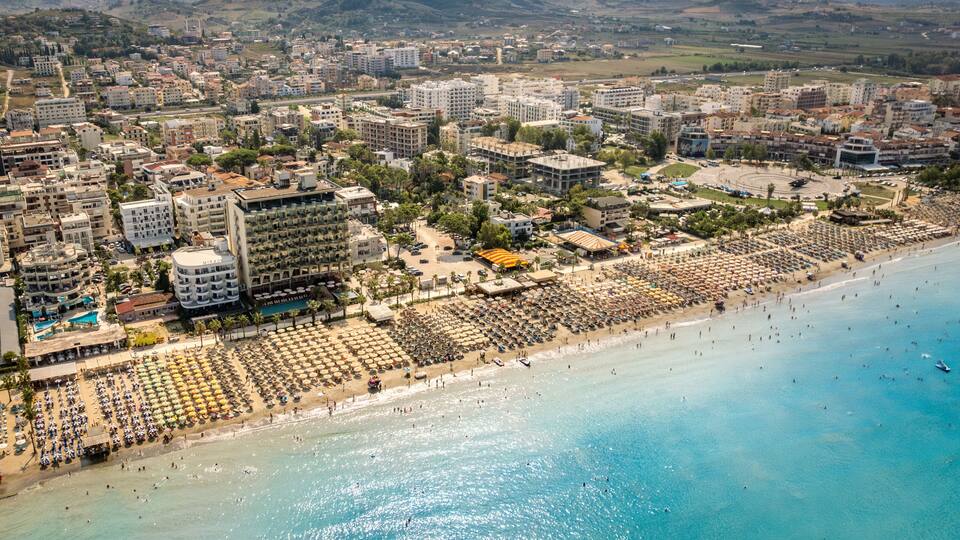 An aerial view of a coastal city with a large beach, numerous umbrellas, and a dense urban area with buildings.