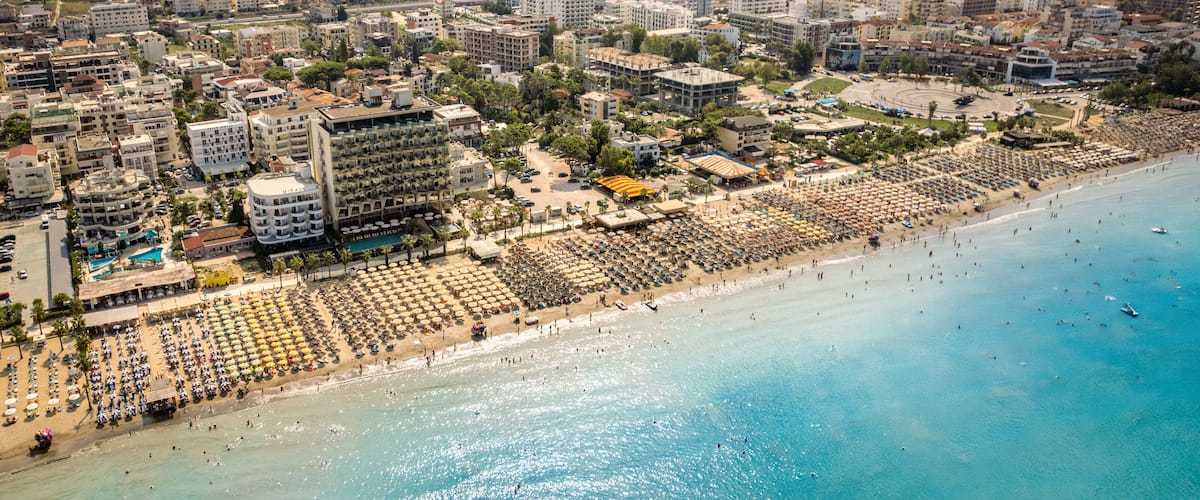 An aerial view of a coastal city with a large beach, numerous umbrellas, and a dense urban area with buildings.