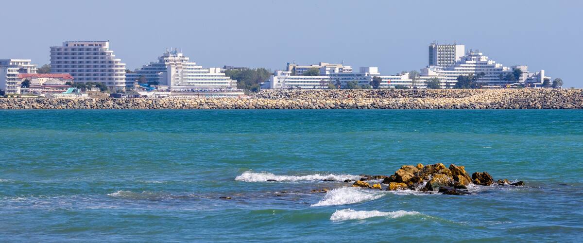 A coastal cityscape with a rocky breakwater and buildings along the shore.