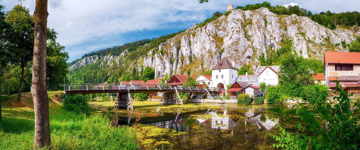 A bridge over a river, surrounded by lush greenery and a rocky mountain in the background.