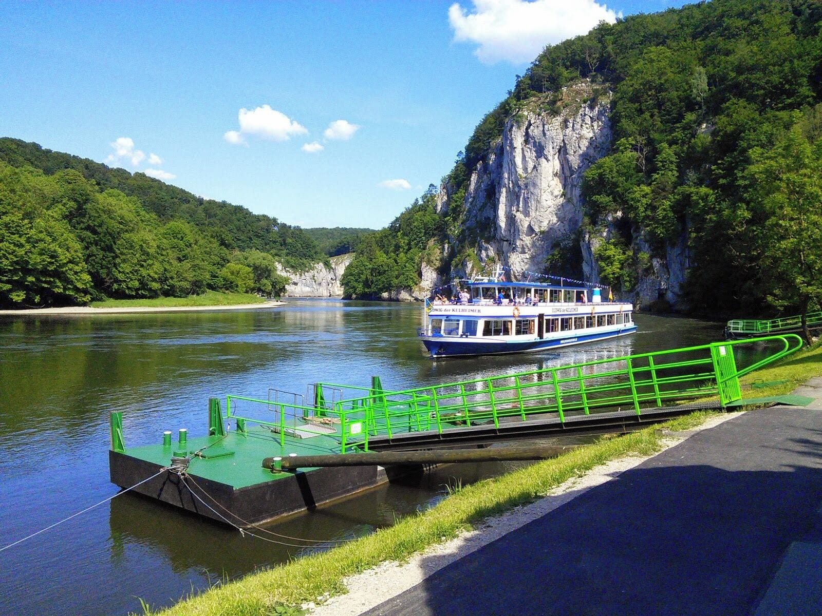 Un río con una barca atracada en un muelle verde, rodeado de colinas verdes exuberantes y un acantilado imponente.