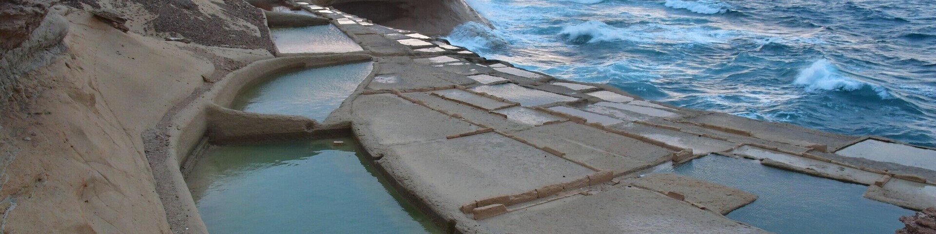 On the north coast of Gozo, just past Qbajjar Bay west of Marsalforn, the coast is characterised by a chequerboard of rock-cut saltpans protruding into the sea. These 350-year-old salt pans, which stretch about 3km along the coast, are more than just scenic. They are part of the centuries-old Gozitan tradition of Sea-Salt production that has been passed down within certain families for many generations. During the summer months, locals can still be seen scraping up the crystals of salt. Once collected, the salt is stored and processed in the caves that have been carved into the coastal rock.