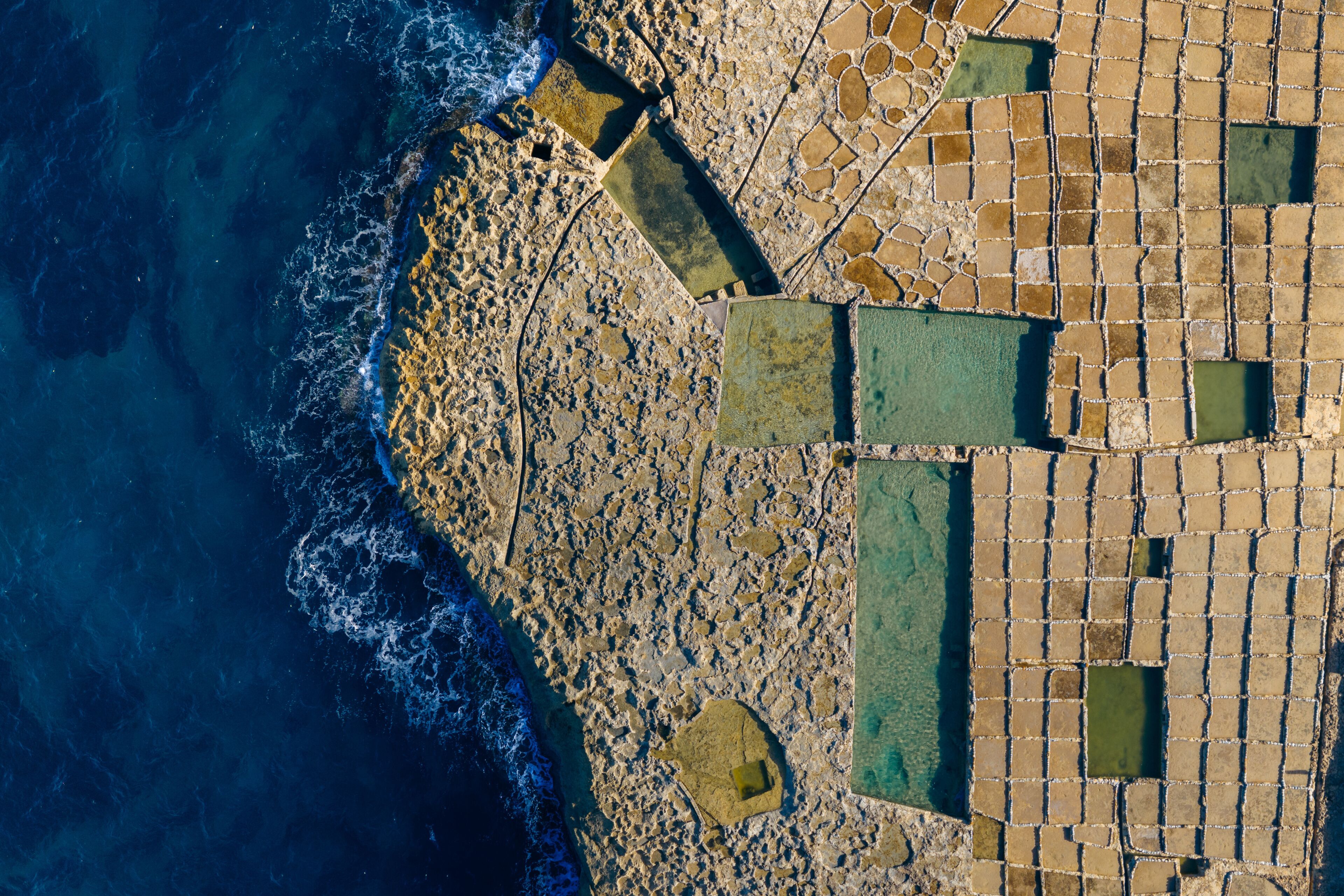 Aerial view of geometric salt pans meet the dark blue sea, creating a stunning contrast of textures and colors, Zebbug, Malta.