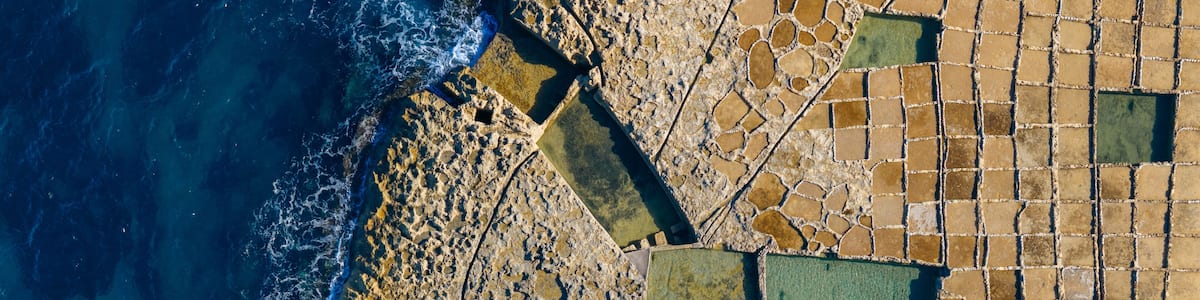 An aerial view of a coastal area with a rocky shoreline, a patterned mosaic pavement, and a clear blue sea.