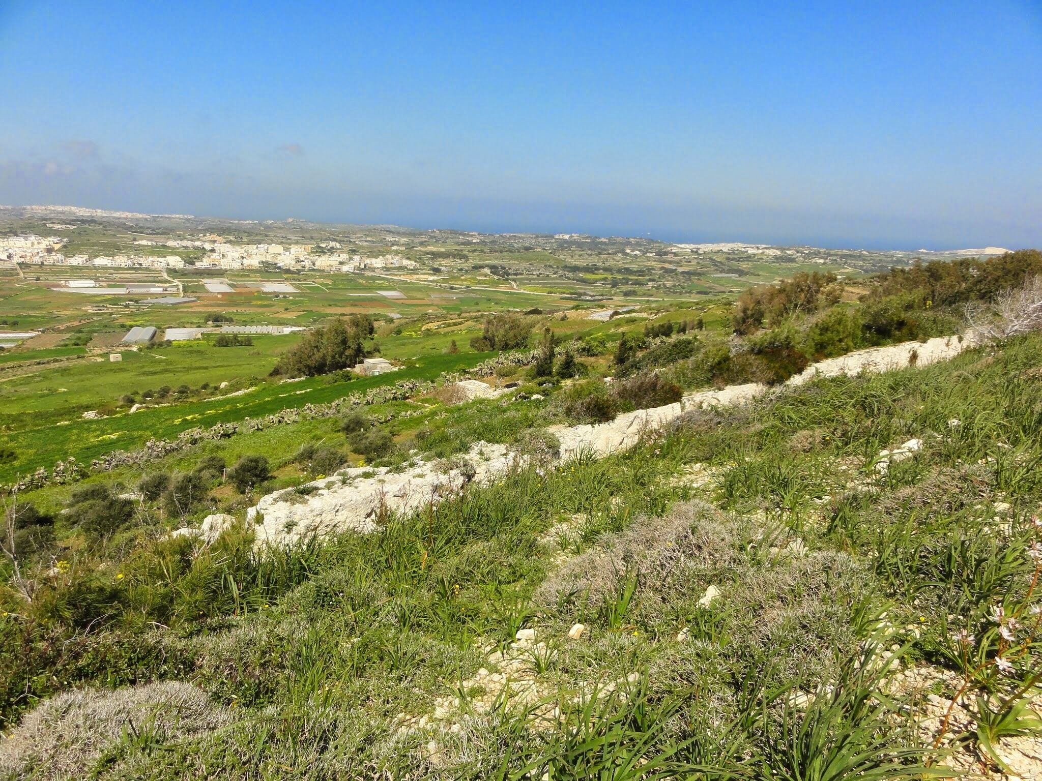 Un paisaje montañoso con campos verdes, edificios dispersos y una vista distante de la costa.