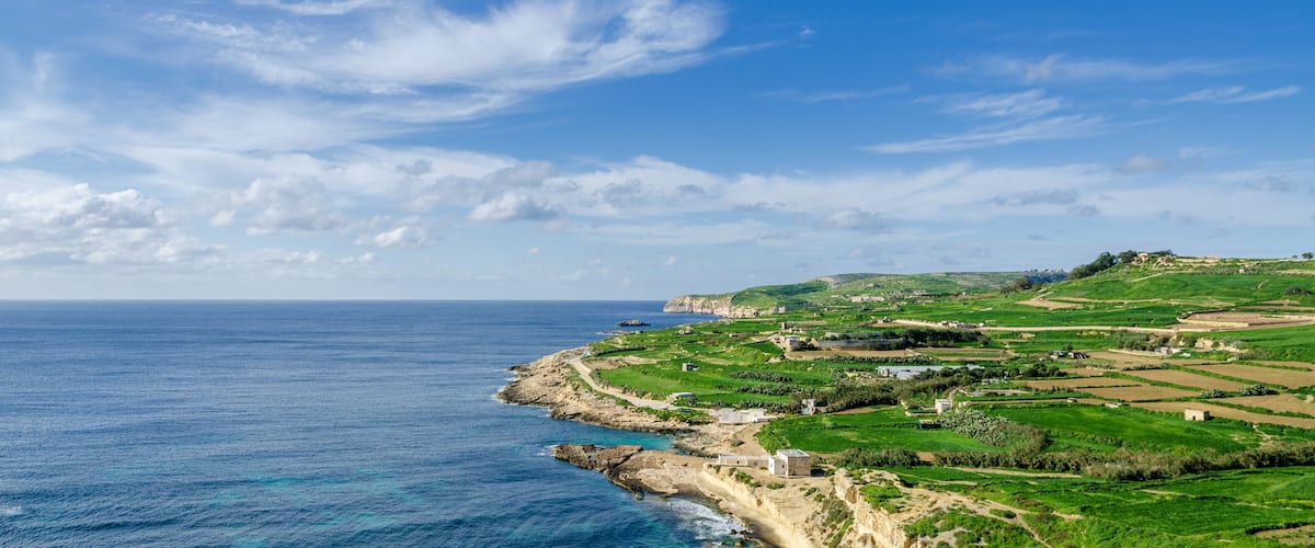 A coastal landscape with a clear sky, rocky coastline, green fields, and scattered buildings.