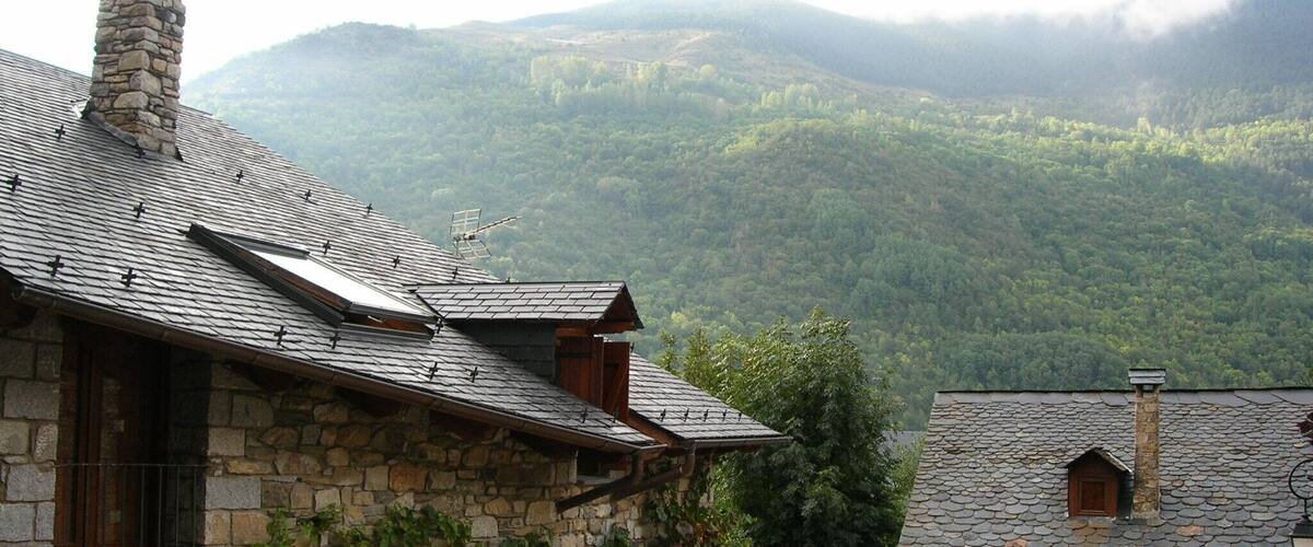A stone building with a sloped roof and wooden shutters, surrounded by lush green hills and a misty mountain backdrop.