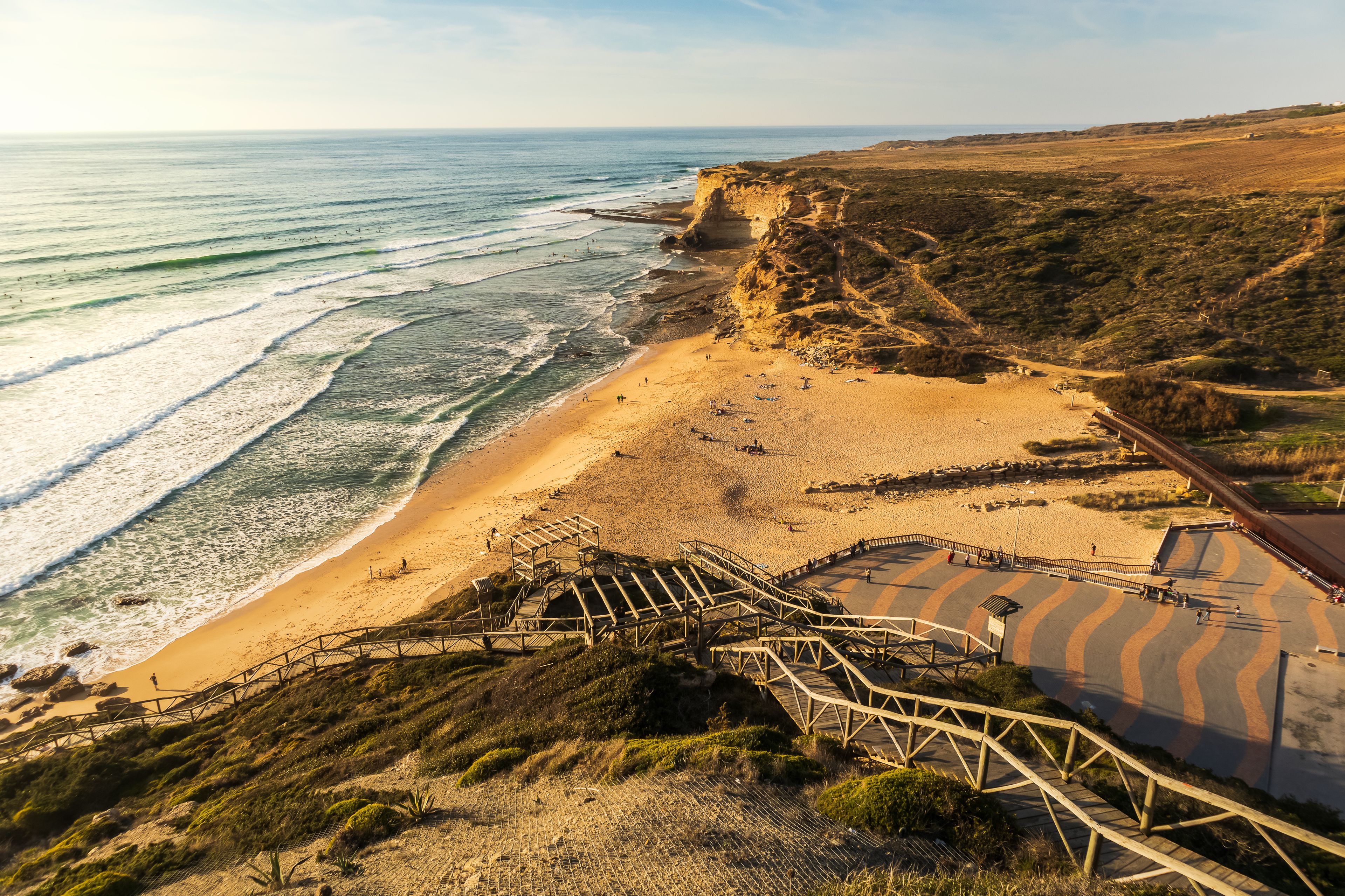Paisagem costeira com praia de areia, ondas do oceano, falésias rochosas e uma escadaria de madeira descendo até a praia.