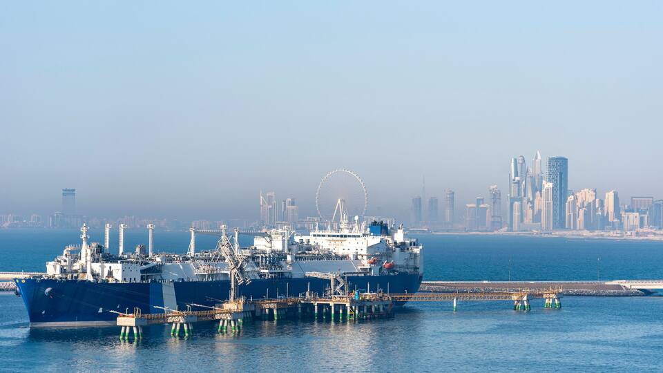 A large cargo ship docked at a port with a city skyline and a Ferris wheel in the background.