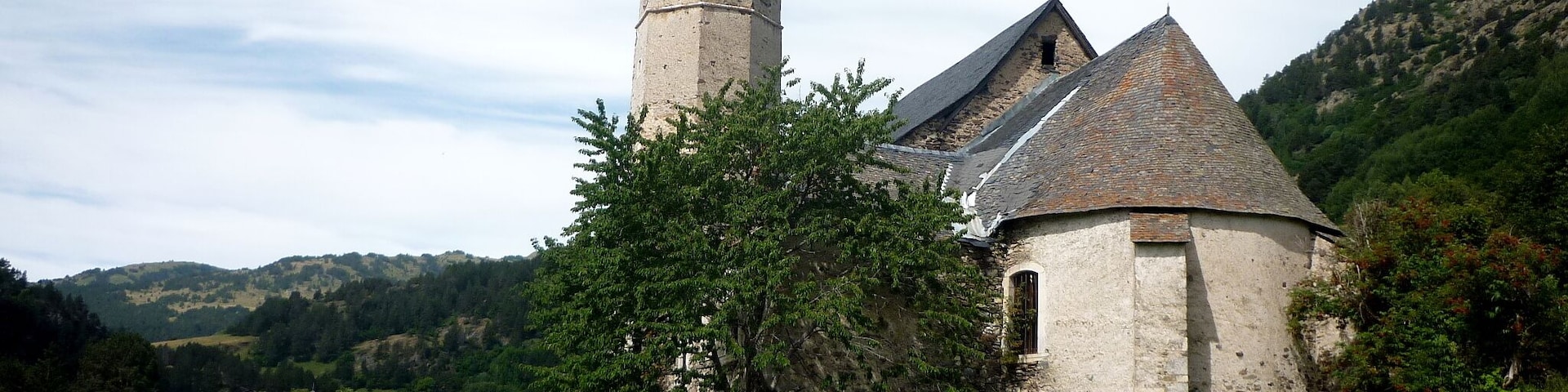 Una iglesia con un alto campanario y un edificio más pequeño, rodeados por un muro de piedra y follaje.