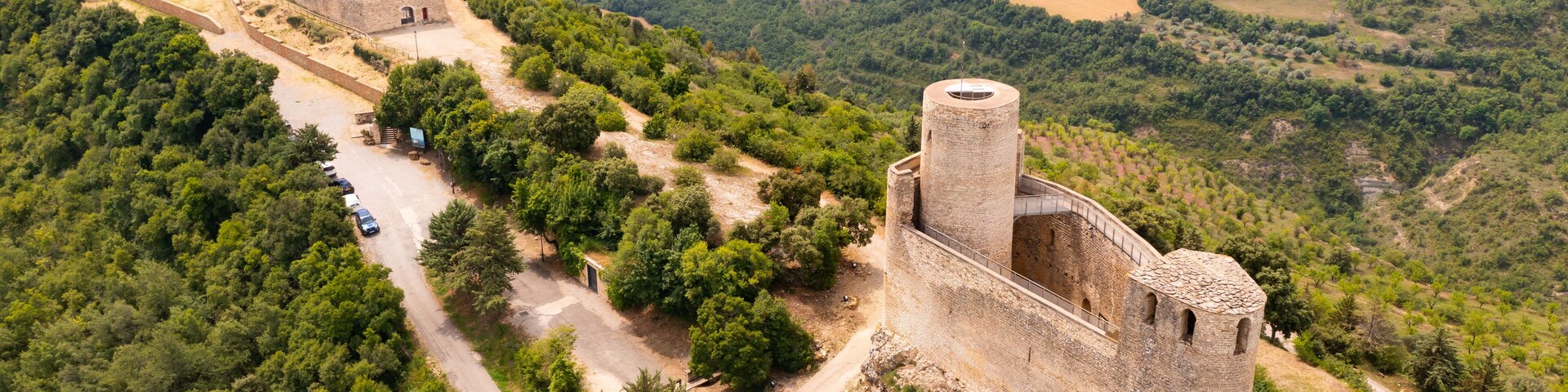Vista aérea de um castelo de pedra histórico cercado por vegetação exuberante e uma estrada sinuosa.