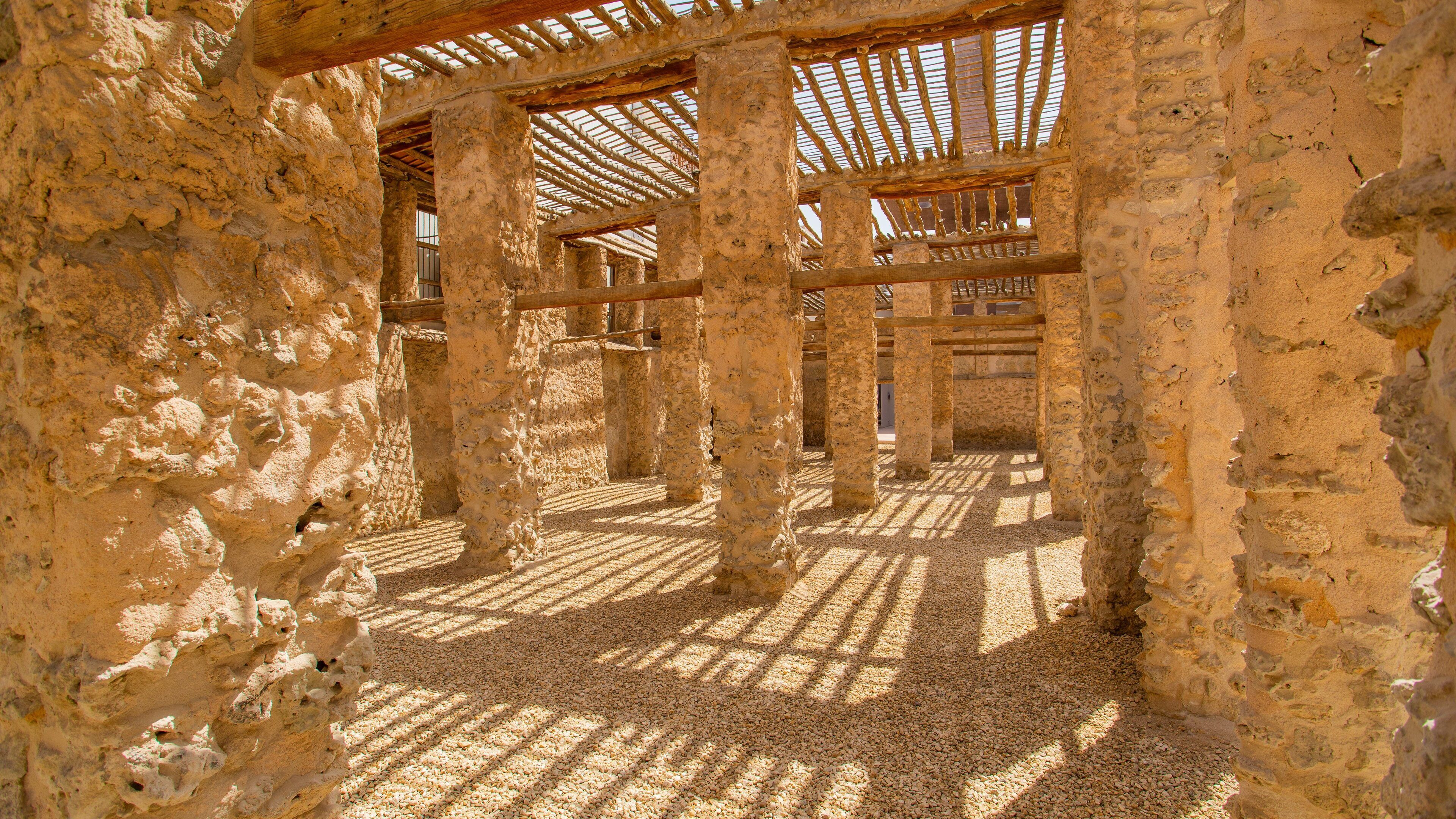 An ancient stone structure with wooden beams and columns, featuring a gravel floor and sunlight filtering through.