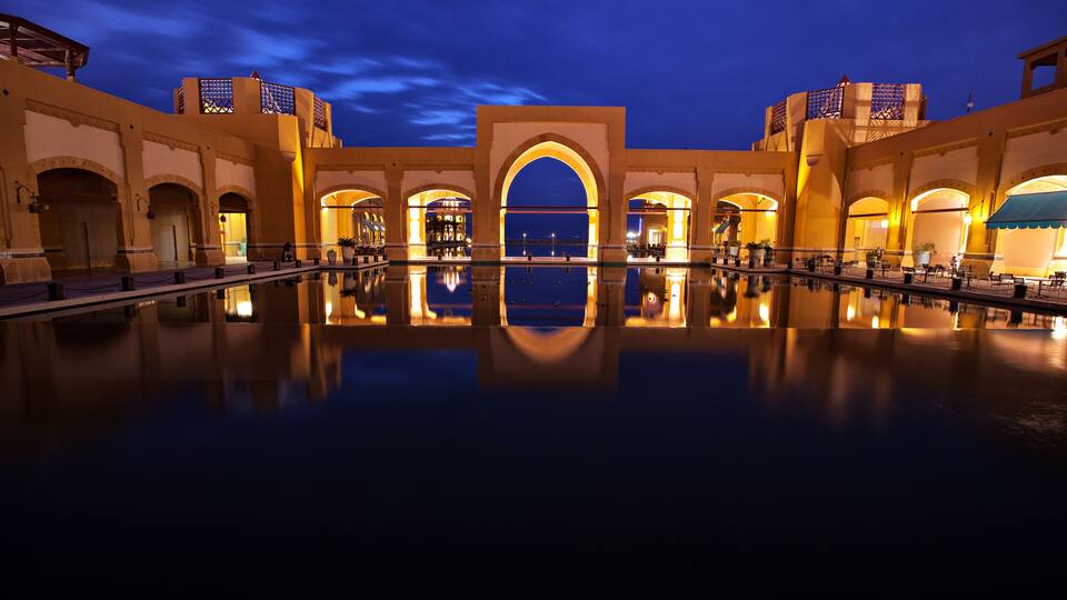 A nighttime view of a luxurious hotel with a large reflecting pool and arched architecture.