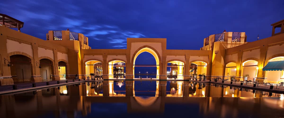 A nighttime view of a luxurious hotel with a large reflecting pool and arched architecture.