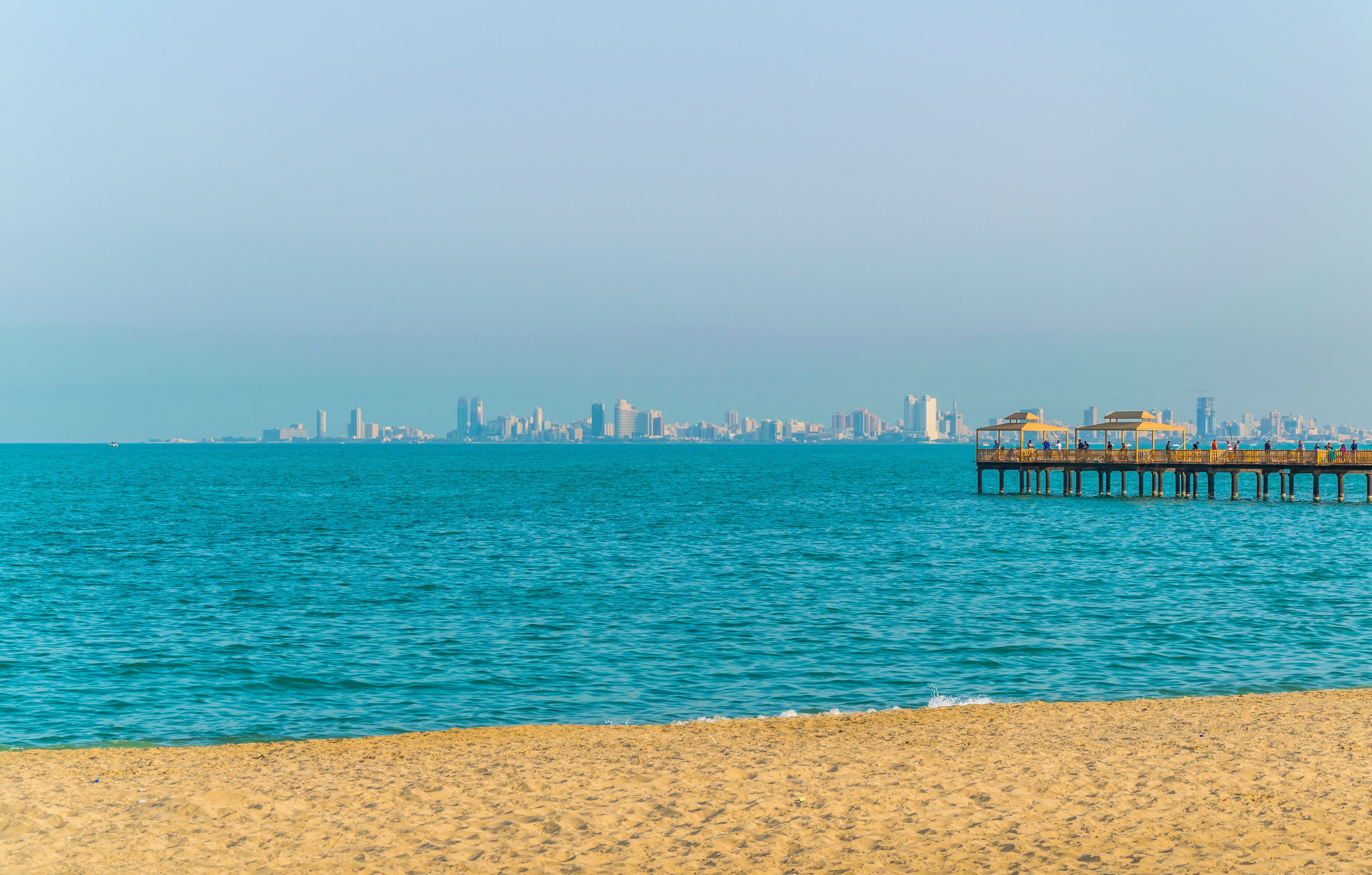 A beach with a pier extending into the water, a city skyline in the background, and a clear sky.
