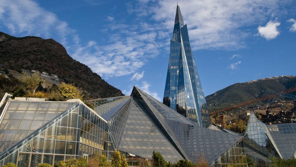 A modern glass building with a pointed top, surrounded by smaller glass structures and a mountainous backdrop.
