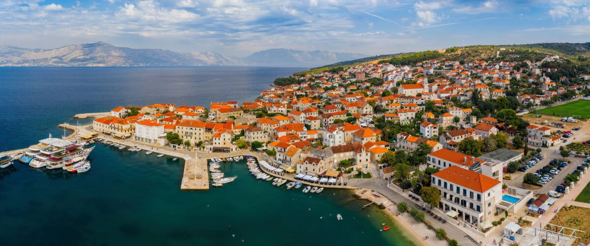 A coastal town with red-roofed buildings, a marina with boats, and a mountainous backdrop.