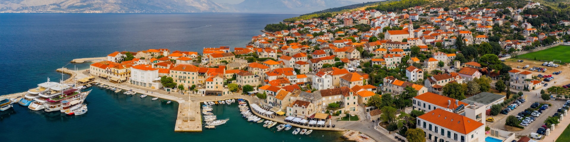 A coastal town with red-roofed buildings, a marina with boats, and a mountainous backdrop.