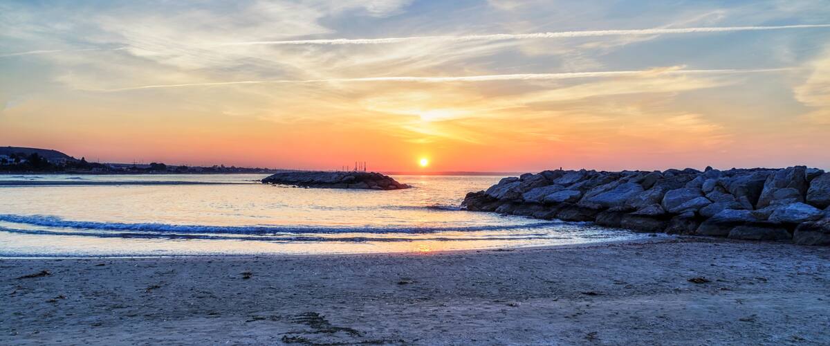 A sunset over a beach with waves, a rocky breakwater, and distant buildings.