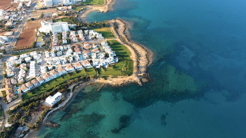 An aerial view of a coastal residential area with clear blue waters, a sandy beach, and a dense cluster of white houses.