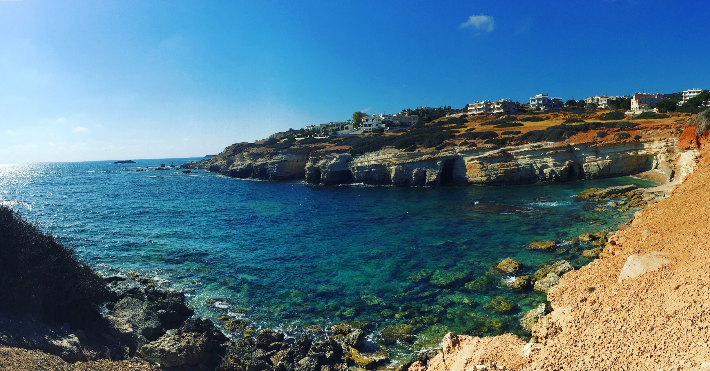 A coastal landscape with clear blue sea, rocky cliffs, and buildings on the hillside.