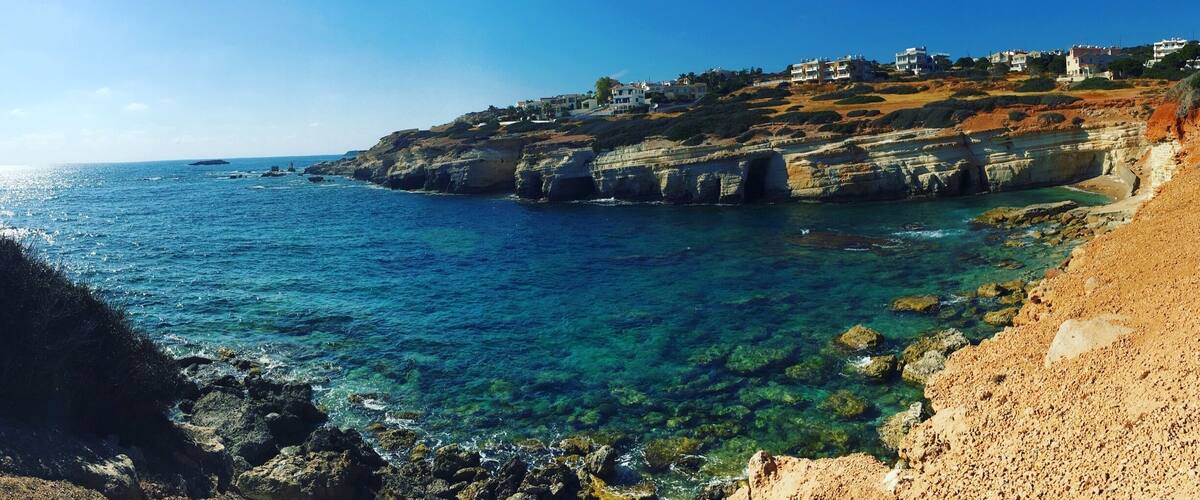 A coastal landscape with clear blue sea, rocky cliffs, and buildings on the hillside.