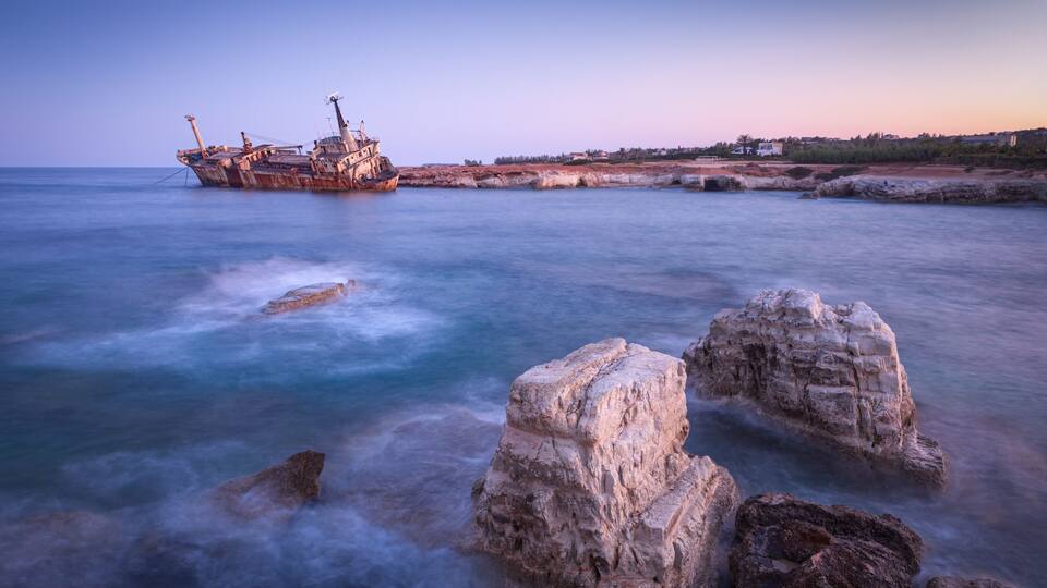 A shipwreck on a rocky shoreline with clear water and a sunset in the background.