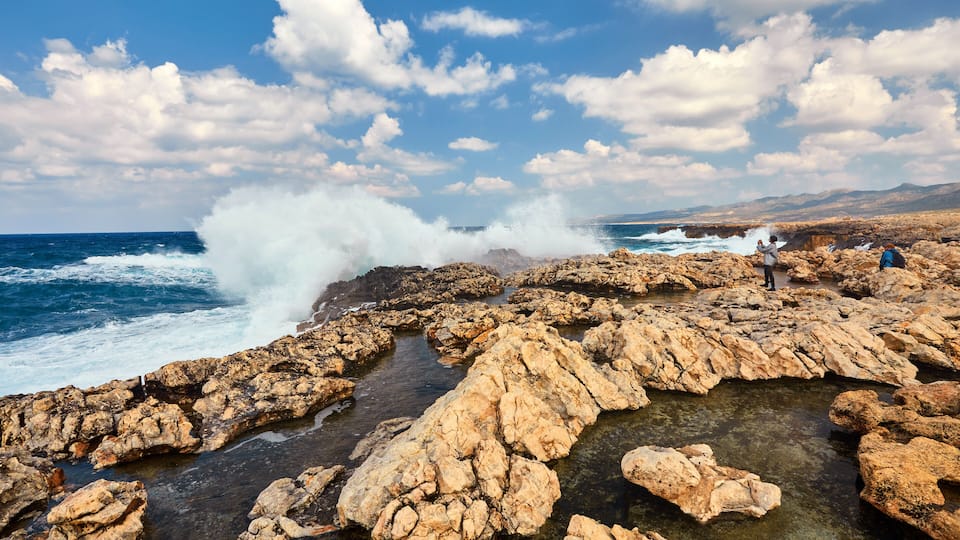 A rocky coastline with waves crashing against the rocks and a clear blue sky.