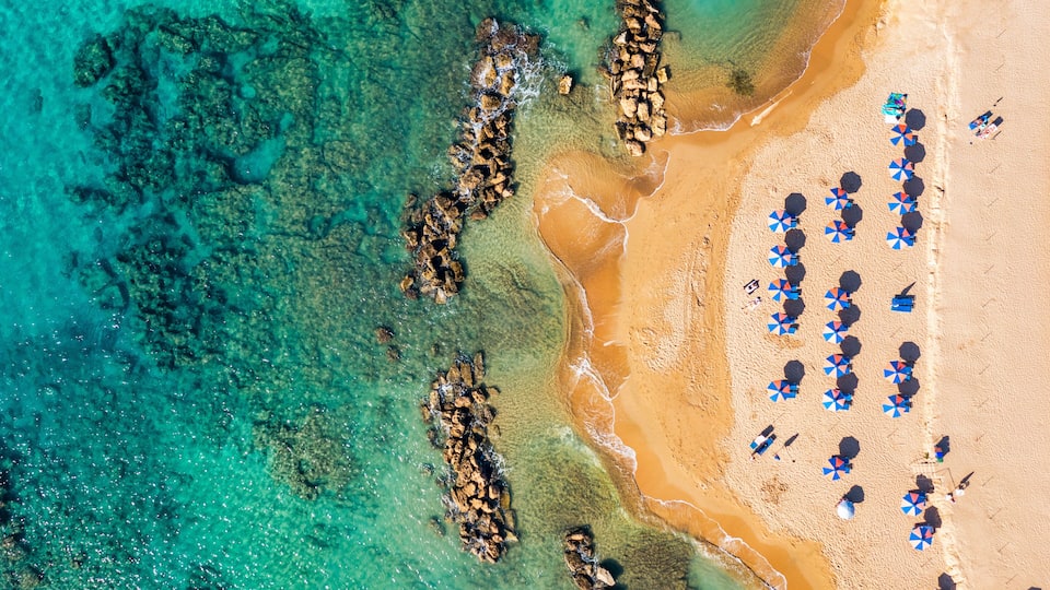 A beach with clear turquoise water, rocky formations, and numerous umbrellas.