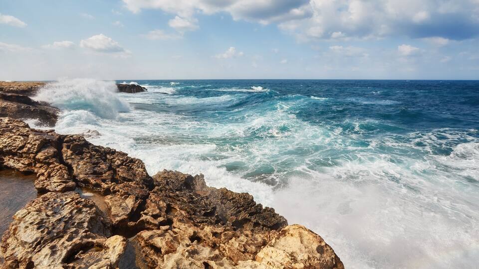 A rocky coastline with waves crashing against the rocks under a partly cloudy sky.