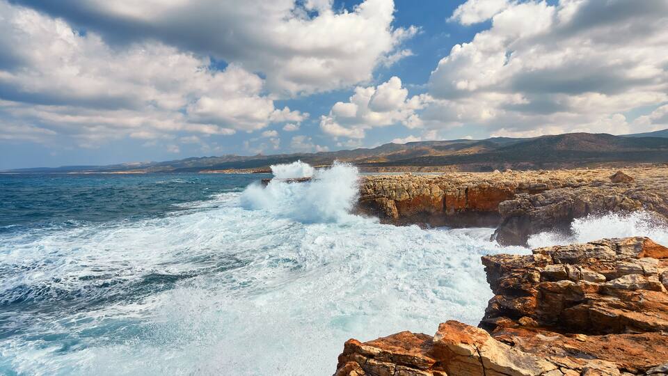 A rocky coastline with waves crashing against the rocks under a partly cloudy sky.