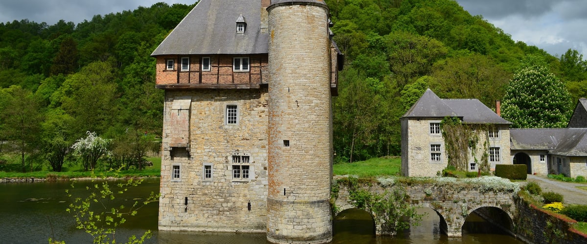 A historic stone castle with a tower, a bridge, and lush green hills in the background.