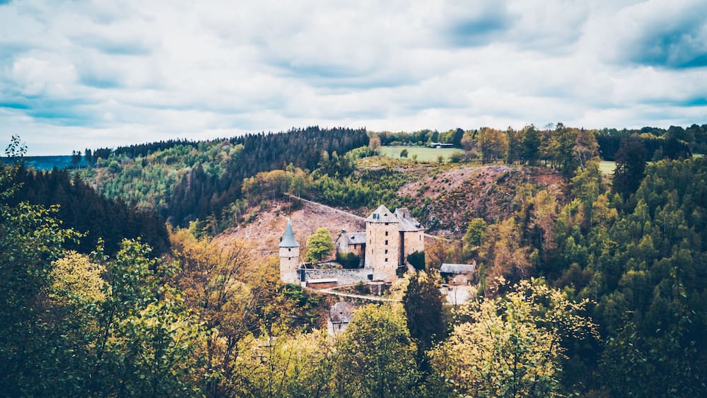 Un château entouré de collines boisées, avec un plan d’eau visible au loin.