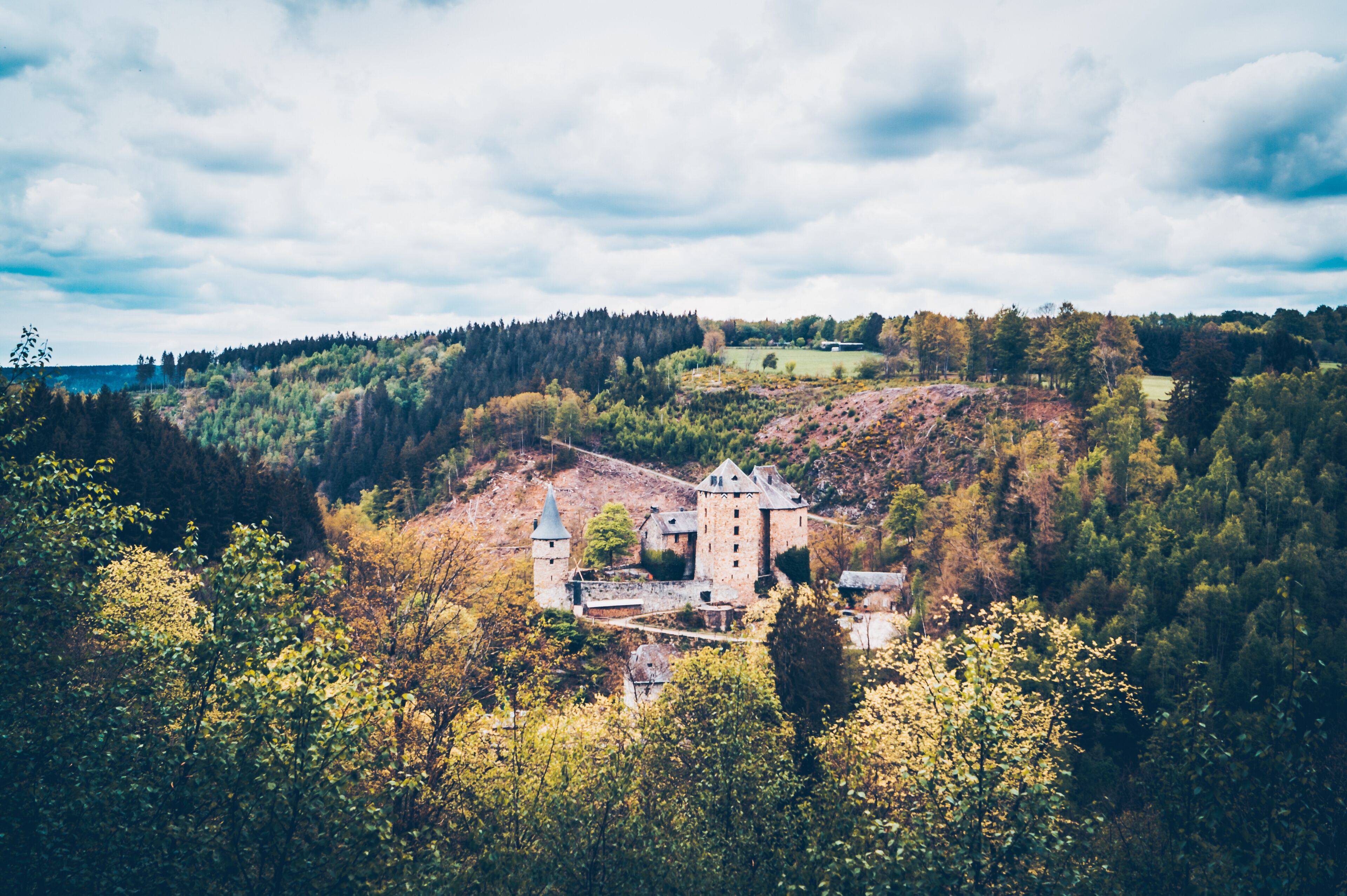 A castle surrounded by forested hills and a body of water in the distance.