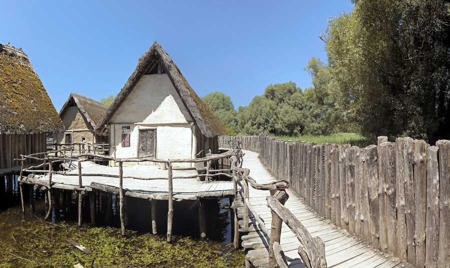 A traditional wooden house with a thatched roof on stilts over water, surrounded by a wooden fence and greenery.