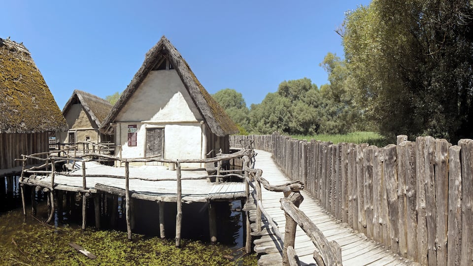 A traditional wooden house with a thatched roof on stilts over water, surrounded by a wooden fence and greenery.
