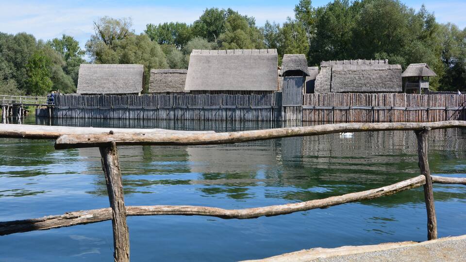 A wooden fence and thatched-roof structures by a calm body of water.