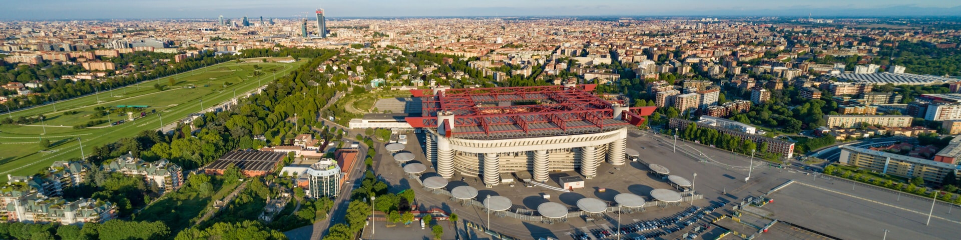 A large circular building with a red roof, surrounded by a parking lot and greenery, in a cityscape with distant buildings and a clear sky.