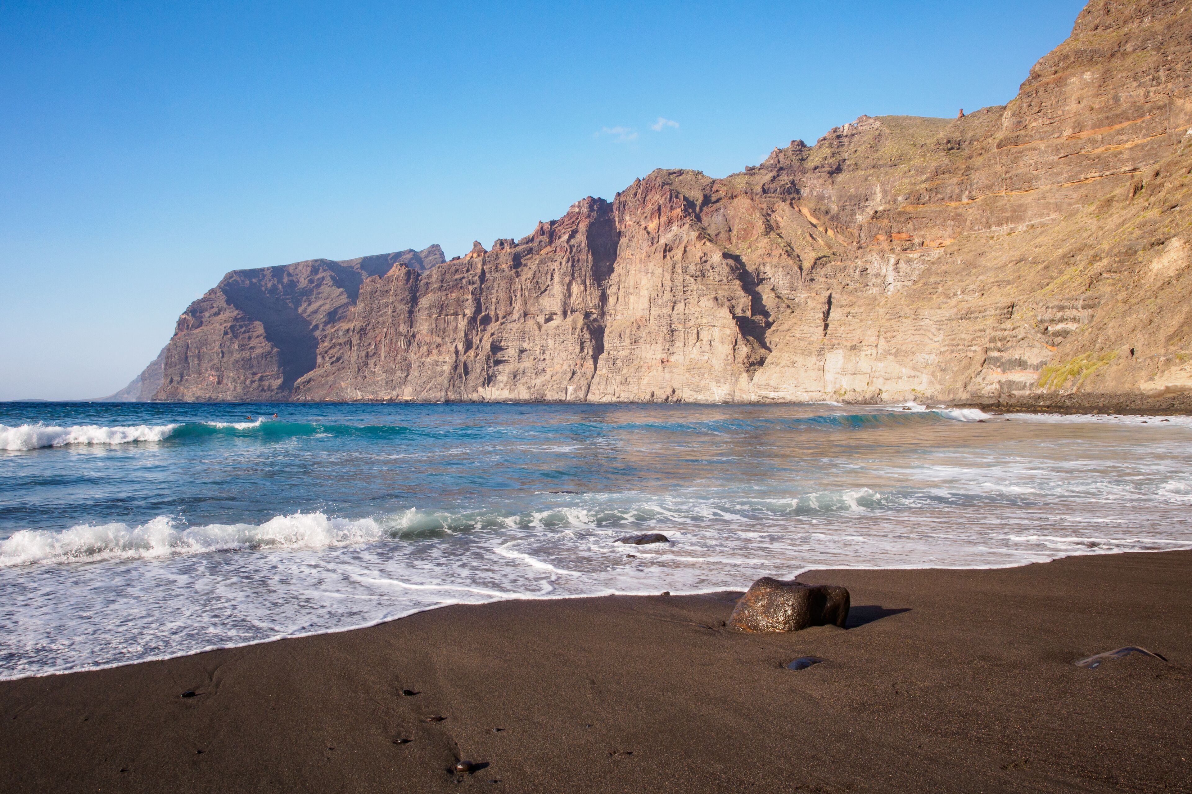 A beach with dark sand, waves crashing onto the shore, and a rocky cliff in the background.