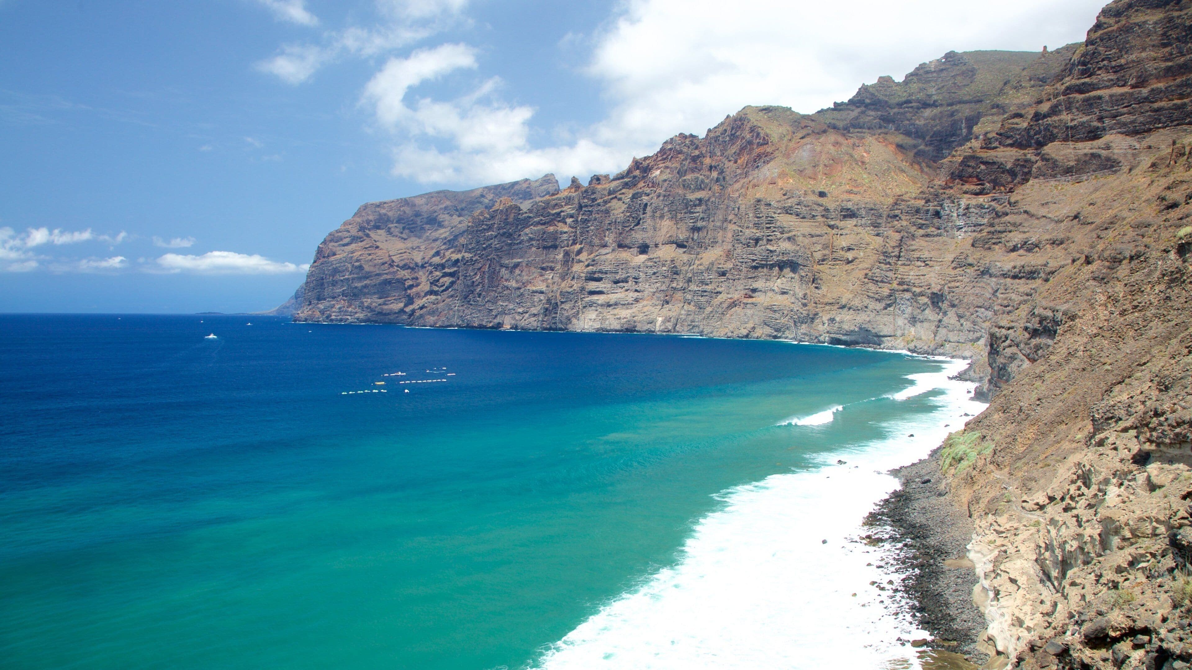 A coastal landscape with a rocky cliff, turquoise sea, and a white sandy beach.