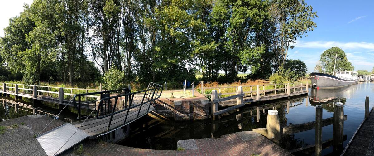 A canal with a wooden bridge, a boat docked on the right, and lush greenery around.