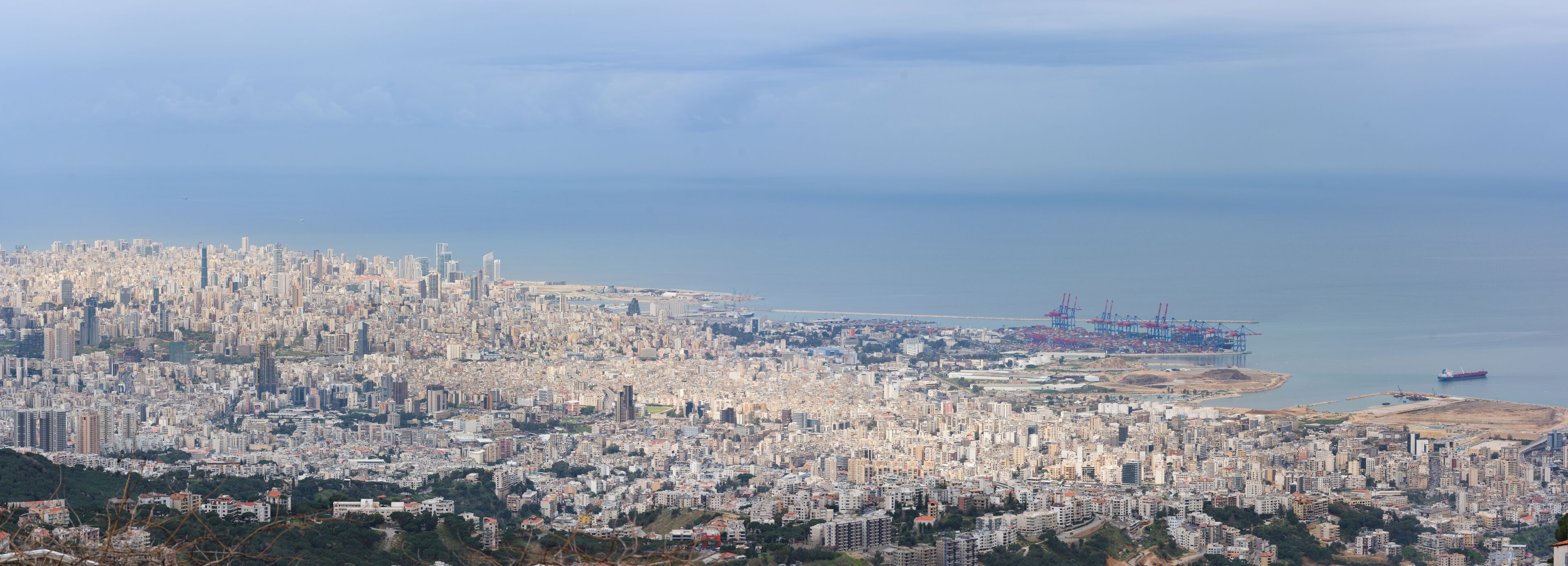 A coastal cityscape with dense urban development, a port with cranes, and a large body of water.