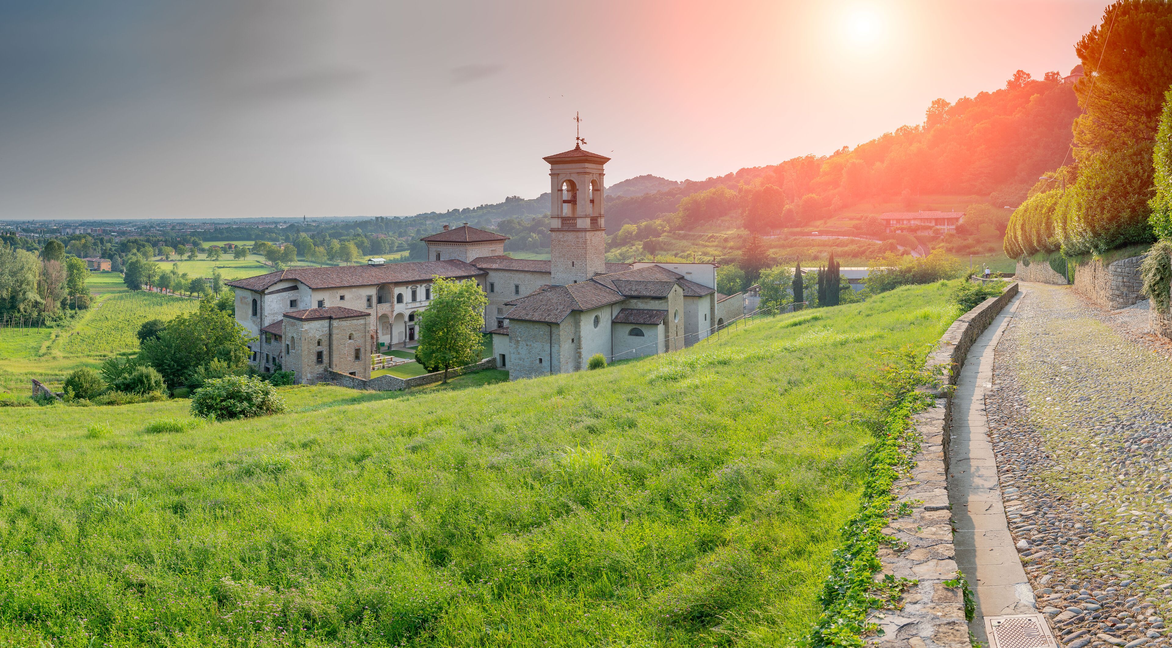 A historic building complex with a bell tower, surrounded by lush green fields and a stone pathway.