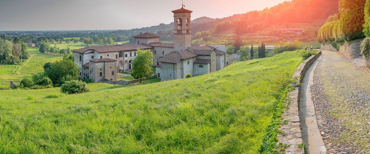 A historic building complex with a bell tower, surrounded by lush green fields and a stone pathway.