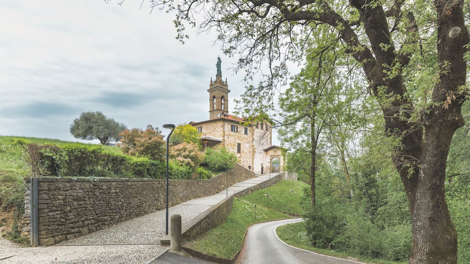 A stone building with a bell tower, surrounded by trees and a gravel path leading to it.