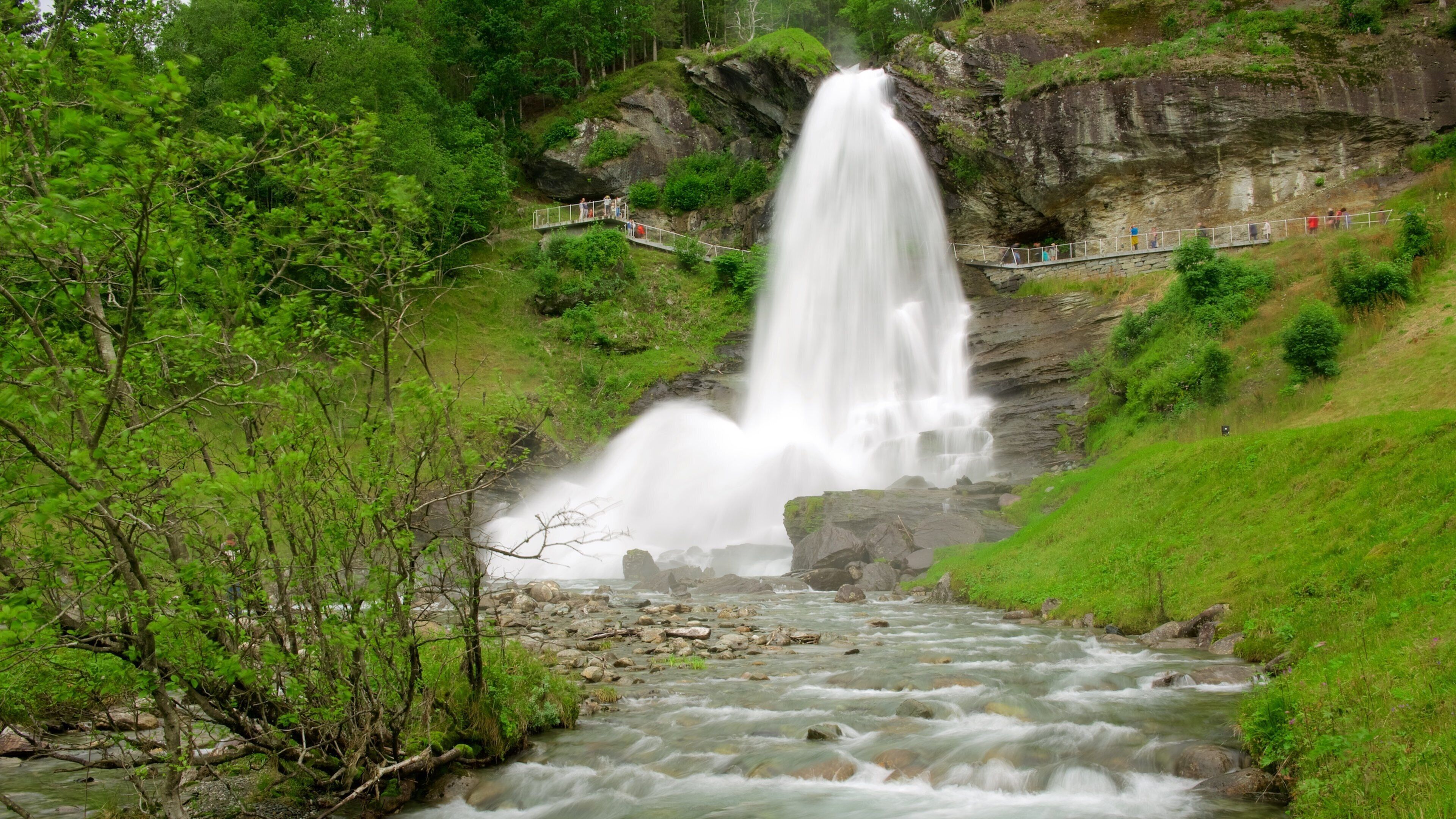 A waterfall cascading down a rocky cliff into a river below, surrounded by lush greenery and a viewing platform.
