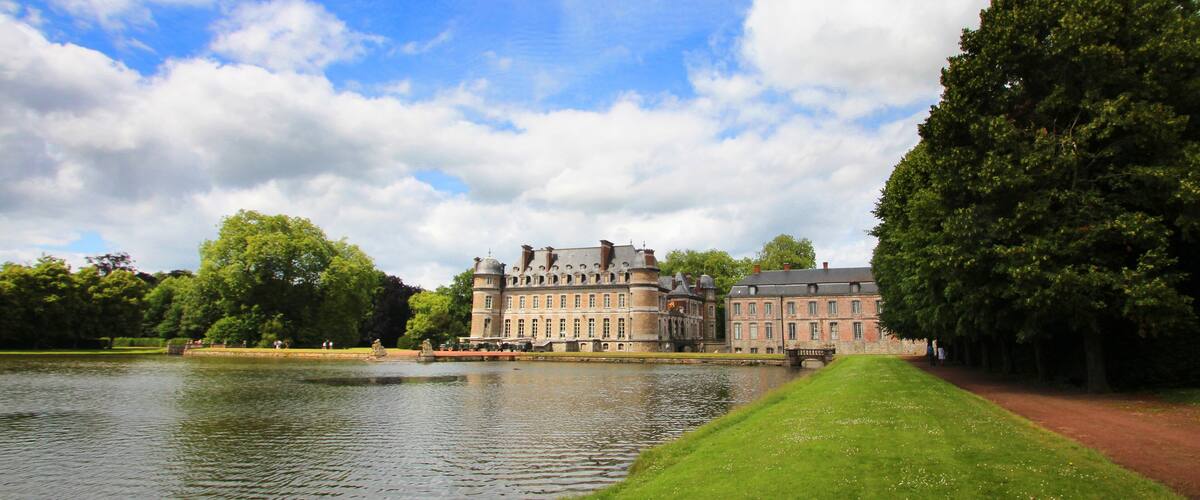 A large historic building with a moat, surrounded by lush greenery and a clear sky.