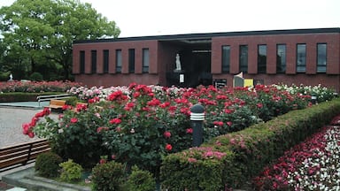 Un bâtiment à la façade en briques rouges, entouré d’un jardin où poussent des fleurs roses et blanches, et orné d’une statue à l’avant.