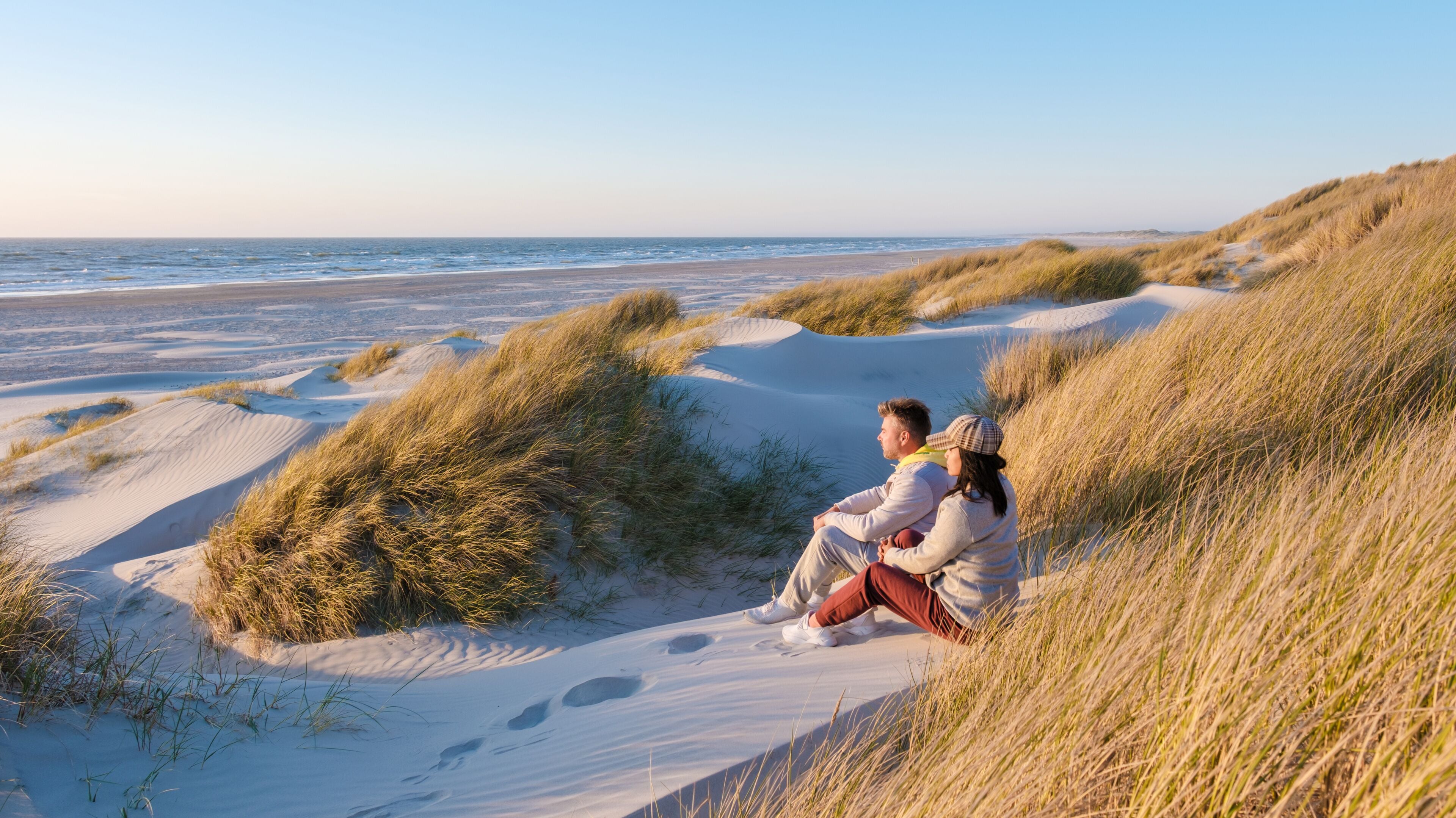 Two people sit together on the soft dunes, surrounded by tall grass, gazing at the calm waves of the North Sea during golden hour in Blokhus Strand,Denmark